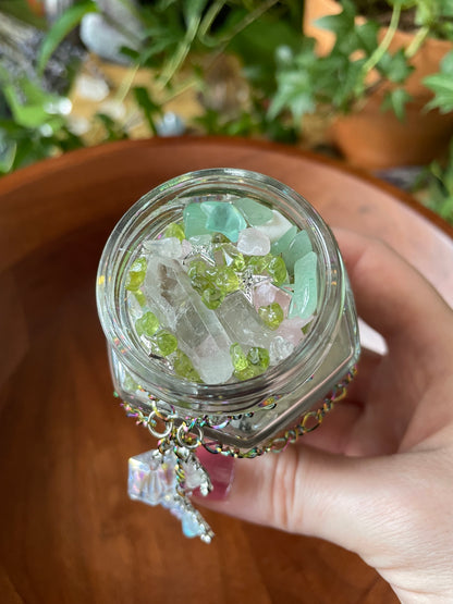 A hand holding a small clear glass jar filled with assorted crystals and gemstones, including pale green, white, and iridescent pieces, with a dangling charm, set against a warm brown wooden background.