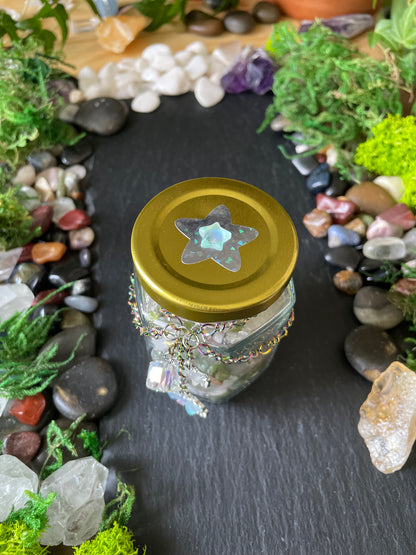 A small clear glass jar with a shiny gold lid and a star-shaped iridescent ornament on top, photographed from above on a dark slate surface surrounded by stones, moss, and crystals.