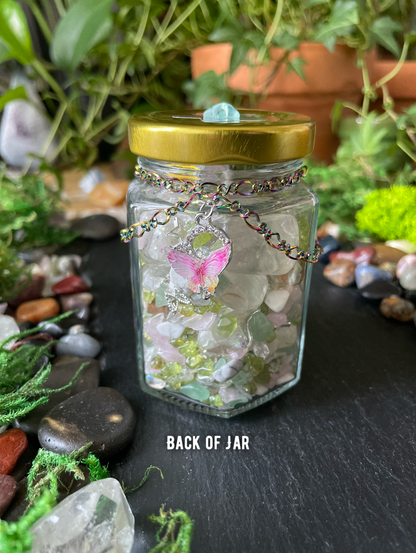 A clear glass jar with a gold metal lid, filled gemstones and decorated with a pink butterfly charm on a chain. The jar is placed on a dark slate surface surrounded by moss, stones, and plants.