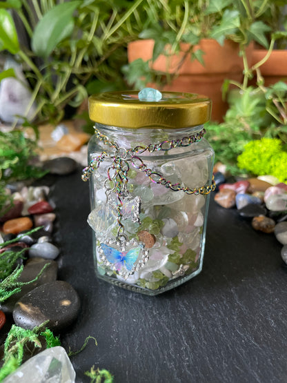 A small clear glass jar with a gold metal lid, filled with gemstones and a silver-toned chain necklace, resting on a dark slate surface surrounded by plants and stones.
