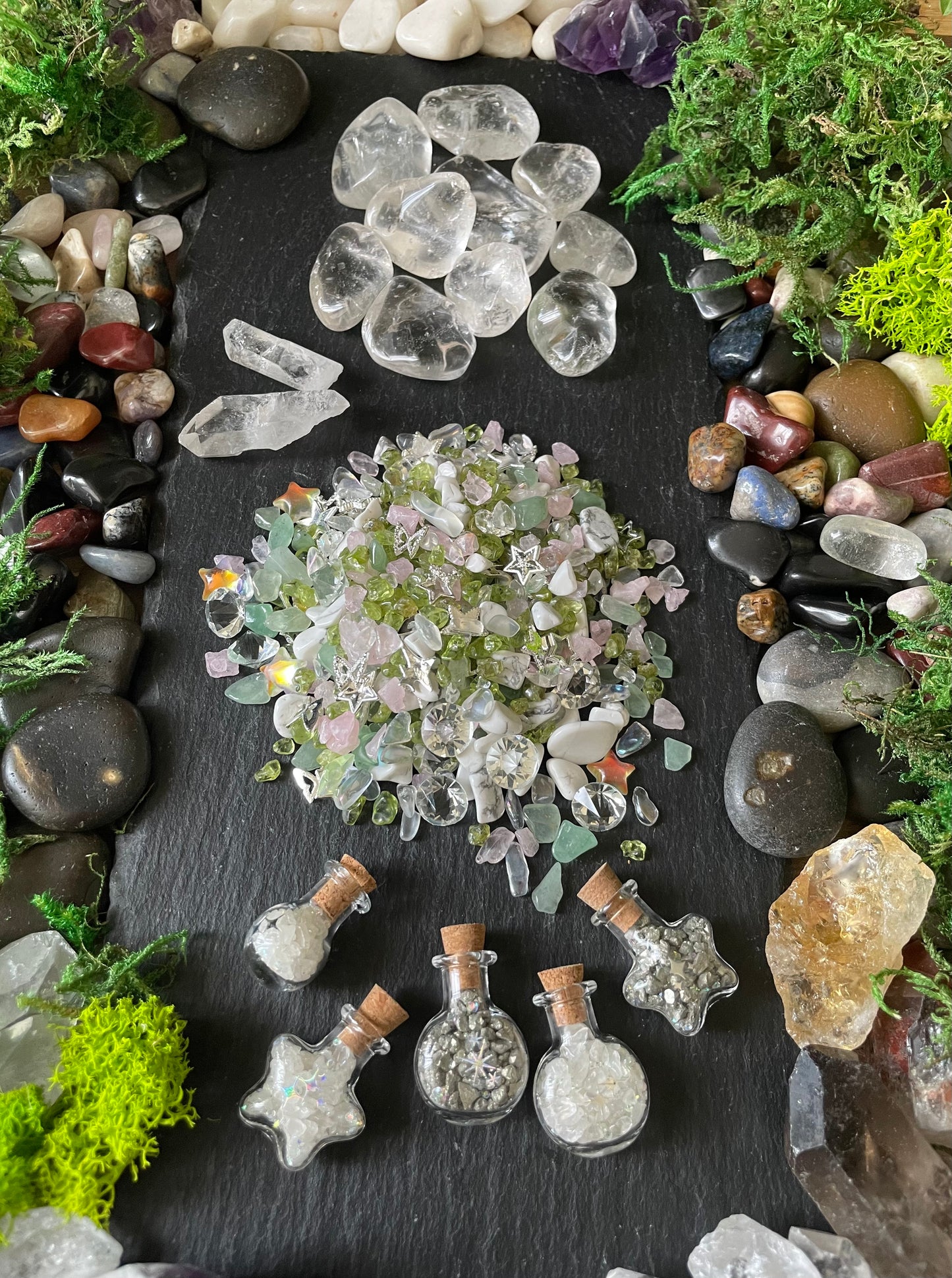 A styled flat-lay of a crystal and gemstone collection on a dark slate background, surrounded by moss, smooth river stones, and small glass potion bottles filled with crystals.