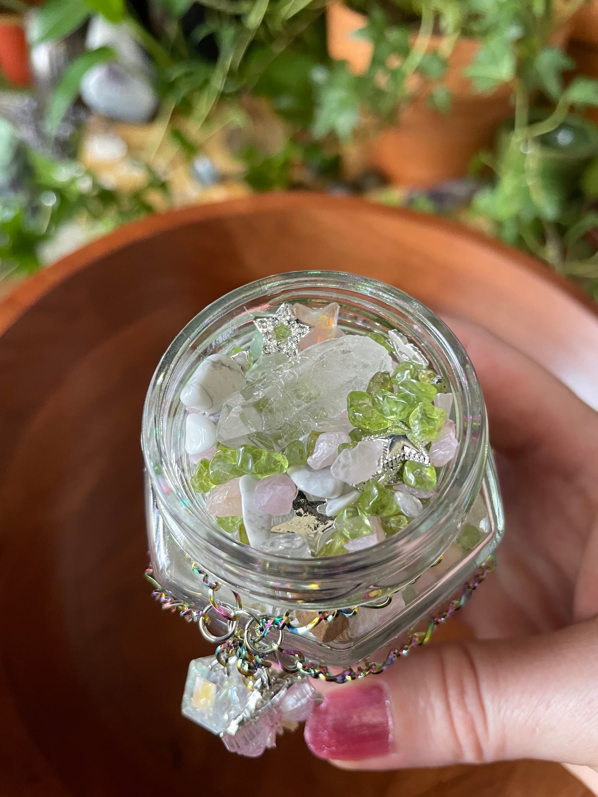 A hand holds a small clear glass jar filled with white and green crystals, with a dangling clear crystal charm, against a blurred background of green plants.