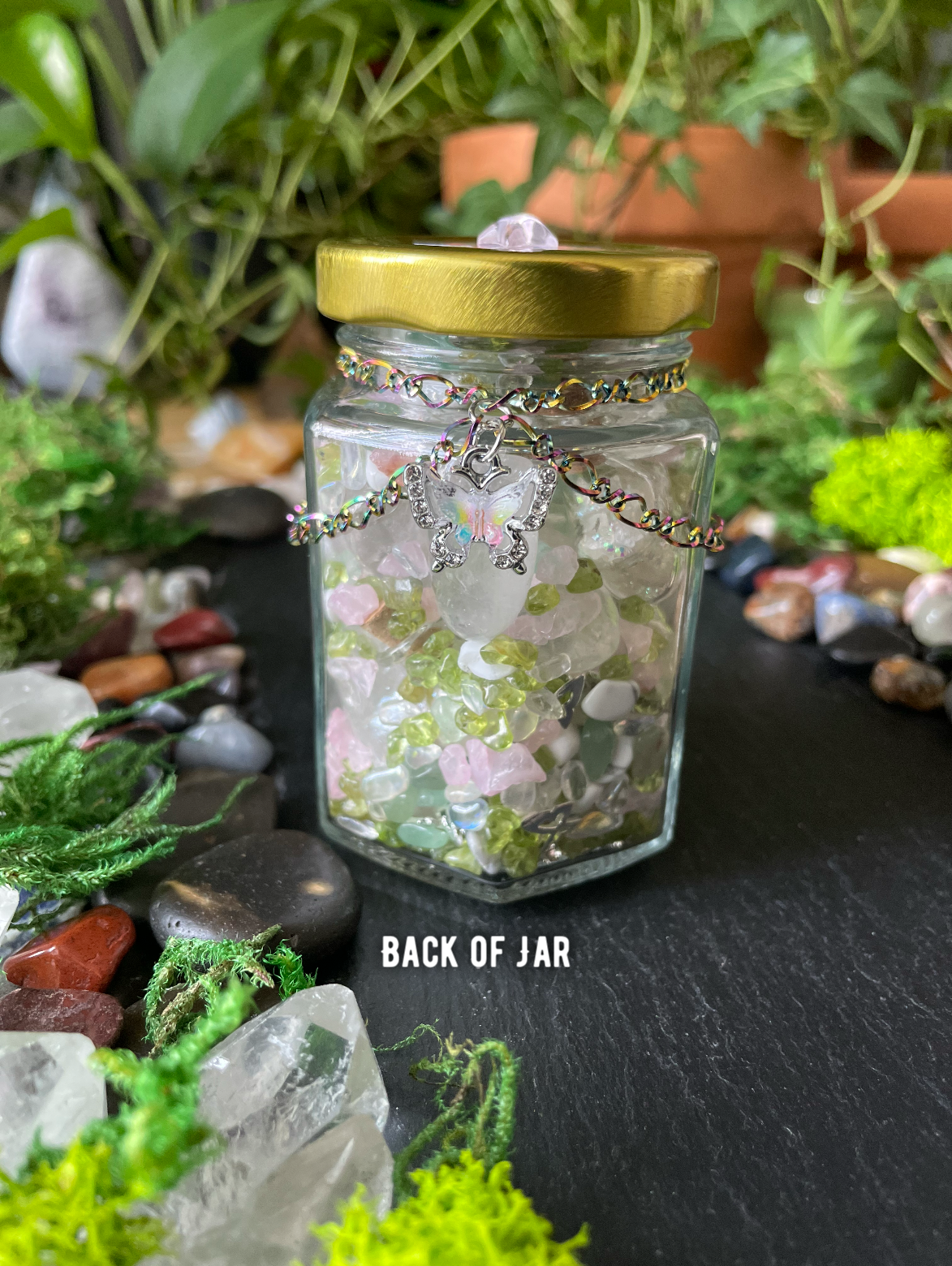 A clear glass jar with a gold metal lid, filled with pastel pink and green crystals, decorated with a rainbow chain and charm, set on a dark slate surface surrounded by moss, stones, and plants.