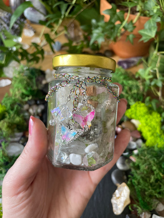 A hand holding a small clear glass jar with a gold metal lid, with pink and iridescent butterfly charms, stones, and star-shaped charms, set against a background of green plants and rocks.