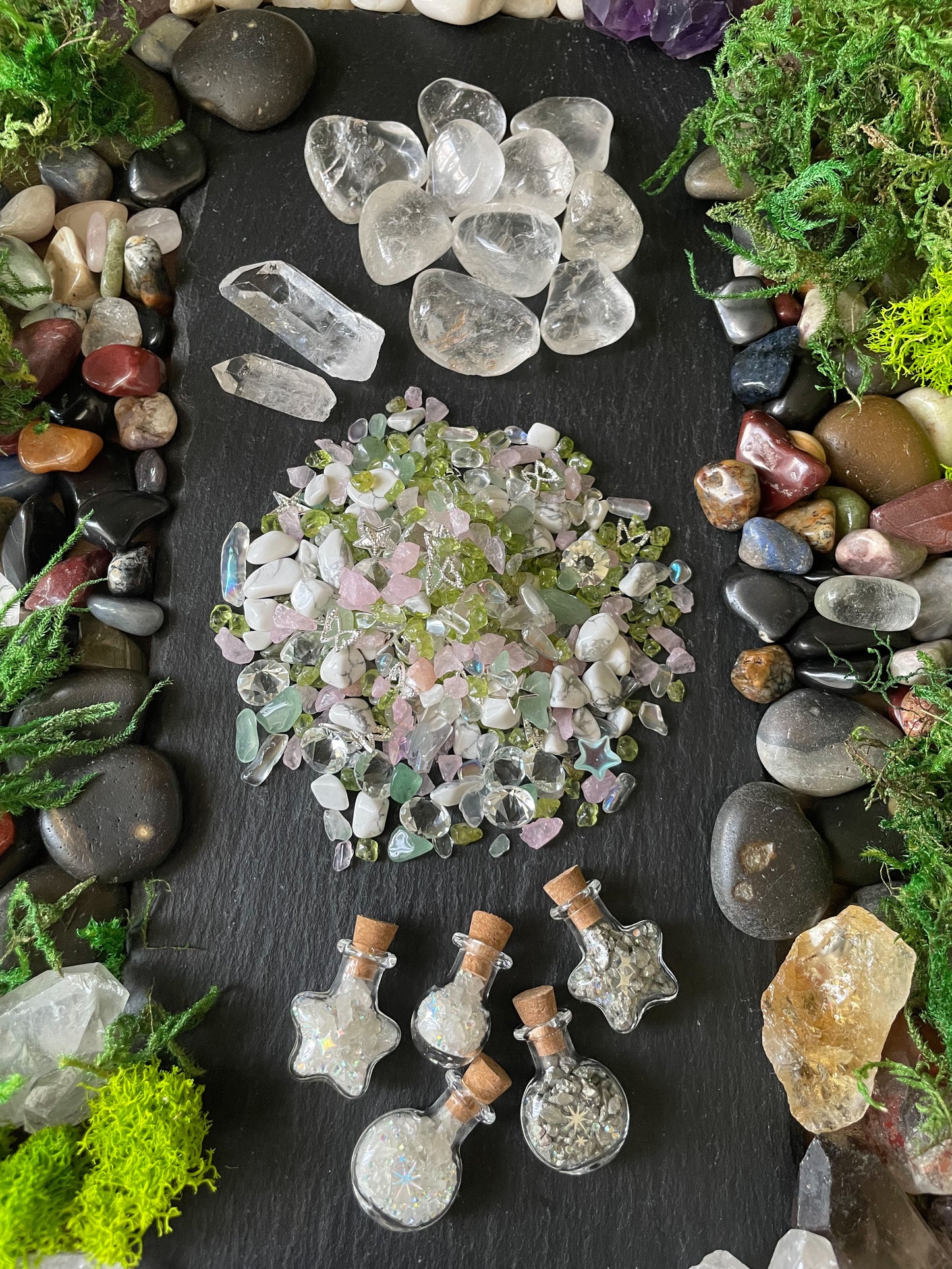 A flat-lay display of various crystals, quartz points, a pile of iridescent pink and green chips, and small glass vials with cork stoppers, all arranged on a dark slate background and framed by natural stones and moss.