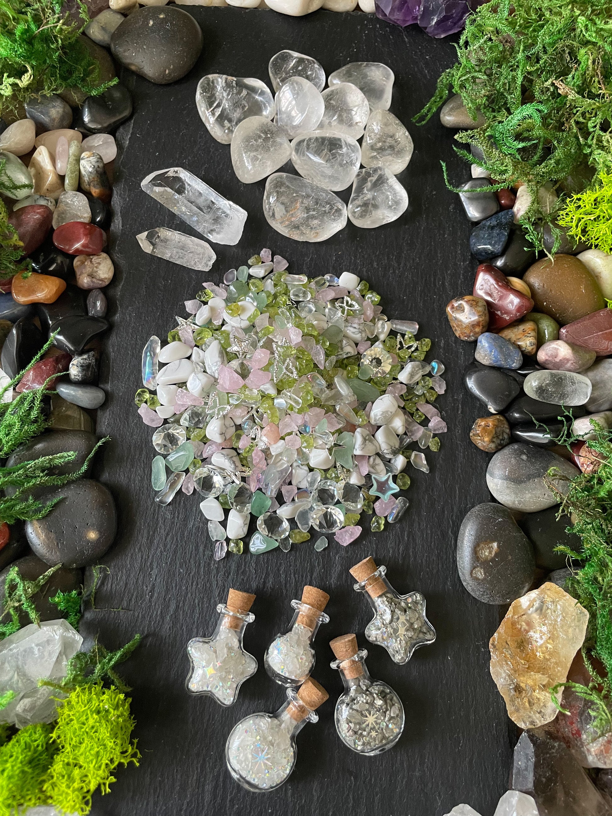 A flat-lay display of various crystals, quartz points, a pile of iridescent pink and green chips, and small glass vials with cork stoppers, all arranged on a dark slate background and framed by natural stones and moss.