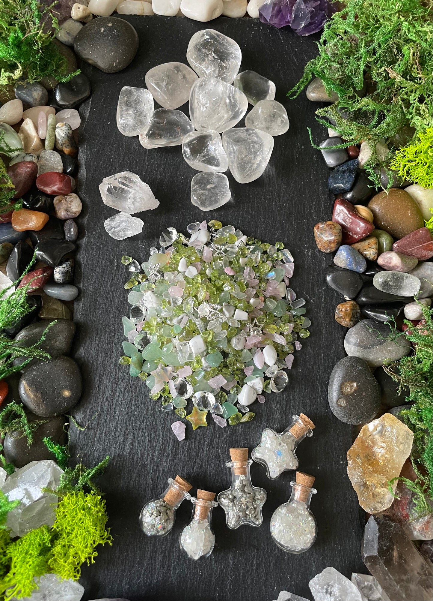 A flat-lay arrangement of various crystals, quartz stones, moss, and small glass vials on a dark slate background.