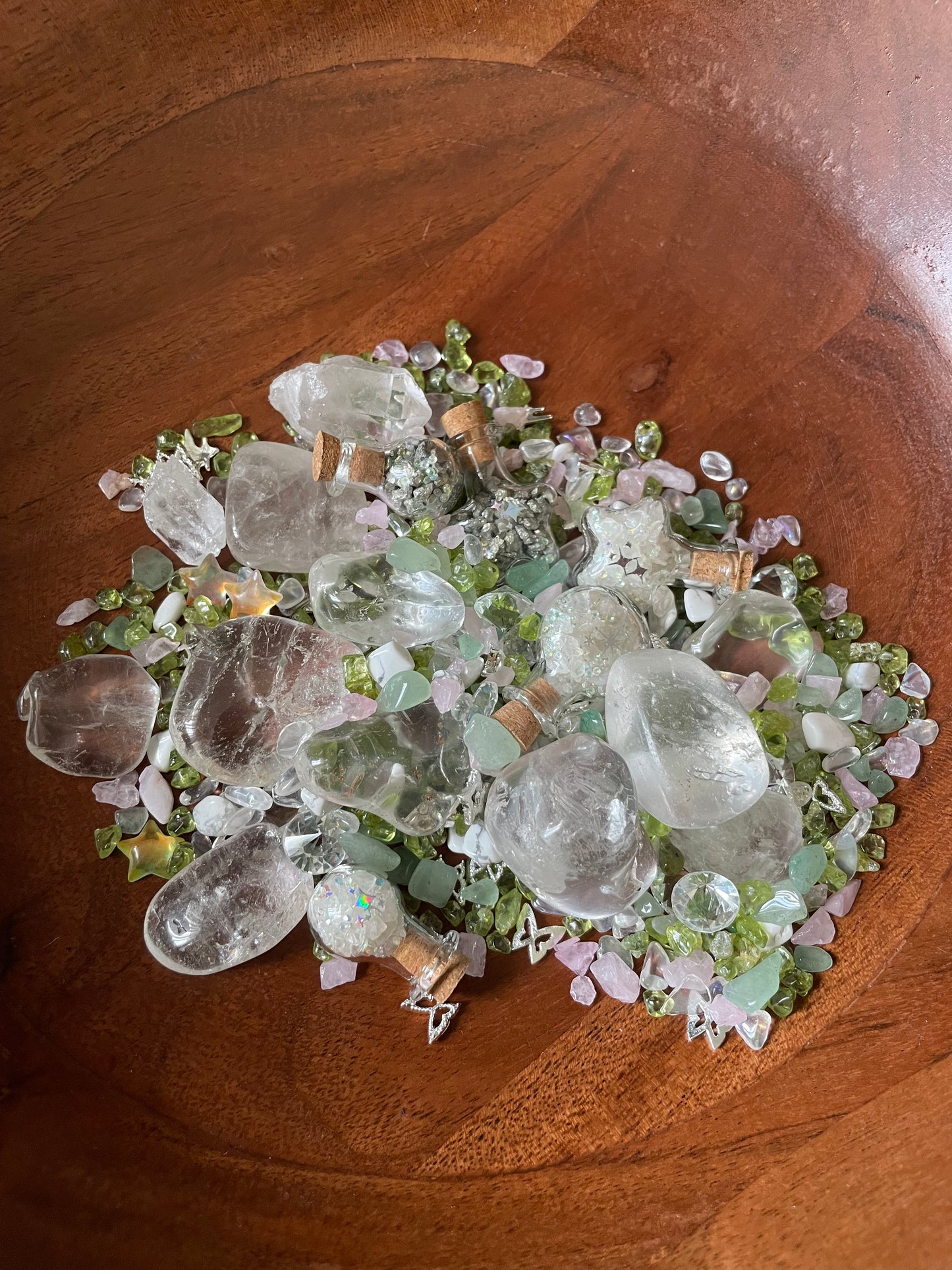 A pile of clear quartz crystal tumbled stones and small pale pink and green gems arranged in a wooden bowl.