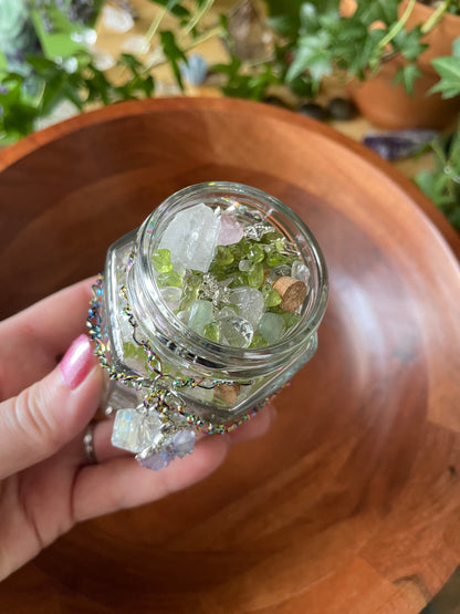 A hand holding a small clear glass jar filled with green and white crystals, set against a wooden bowl background.