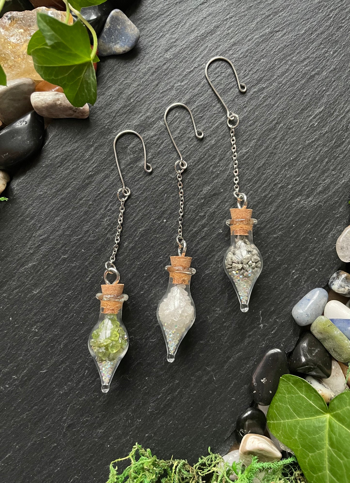 Three small glass vials with cork toppers on a dark slate surface with stones and leaves.