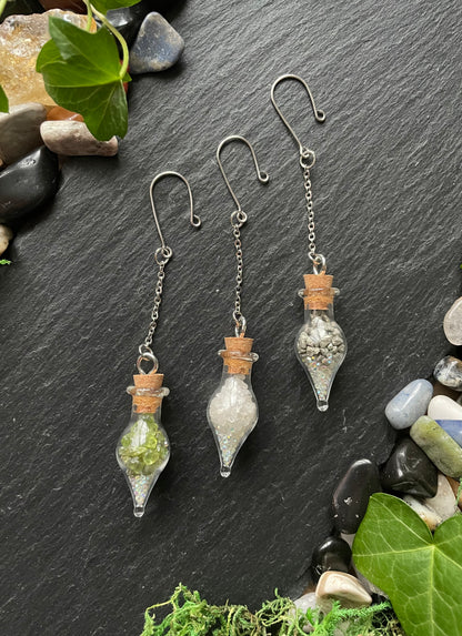 Three small glass vials with cork toppers on a dark slate surface with stones and leaves.