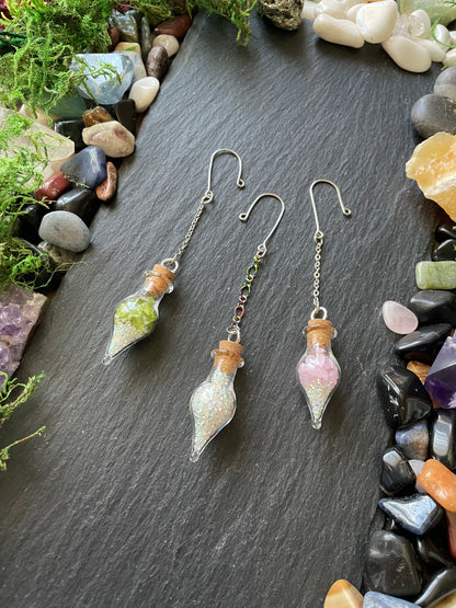 Charms with green, pink, and sparkly decorative gems on a dark slate surface with stones and moss.