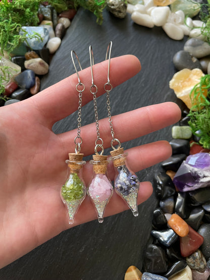 Hand holding three small glass containers with crystals and gemstones on a dark surface with stones and plants.