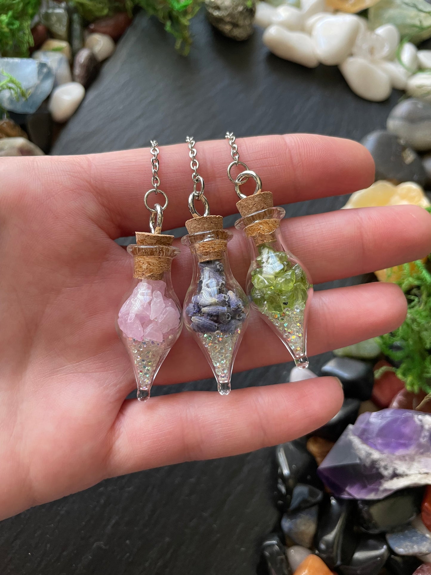 Three small glass bottles with cork lids containing different colored crystals held in a hand against a background of stones and crystals.