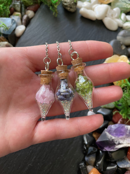Three small glass bottles with cork lids containing different colored crystals held in a hand against a background of stones and crystals.
