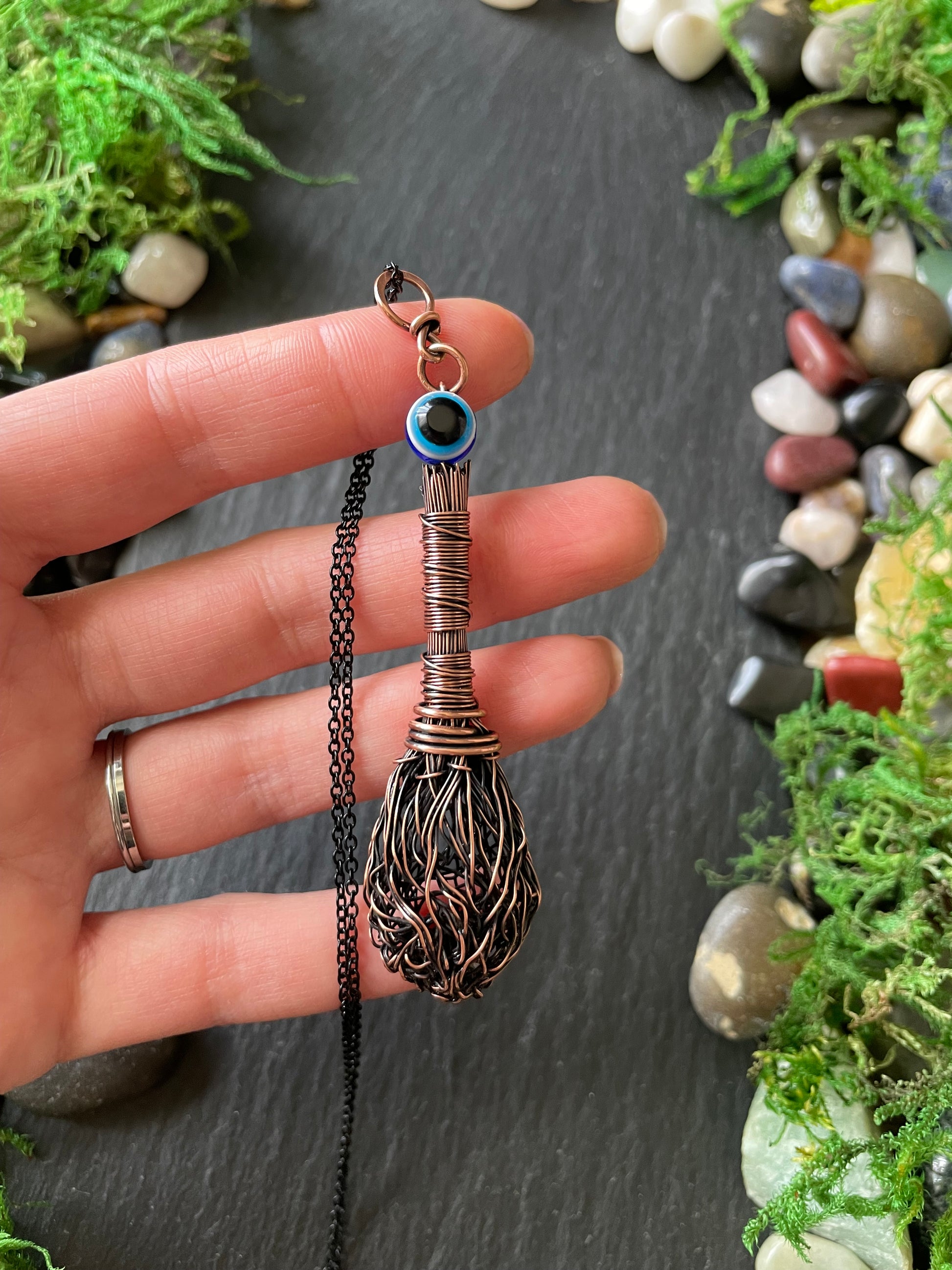 Hand holding a decorative pendant with a blue evil eye bead against a natural stone and moss background