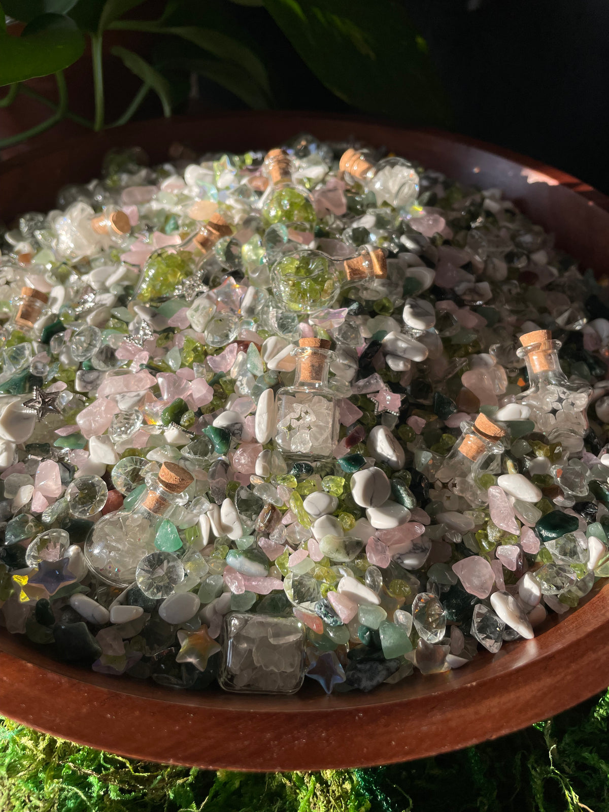 A wooden bowl filled with decorative crystal and stone pieces, including clear glass cubes, pink and green stones, photographed in natural light with dark foliage in the background.