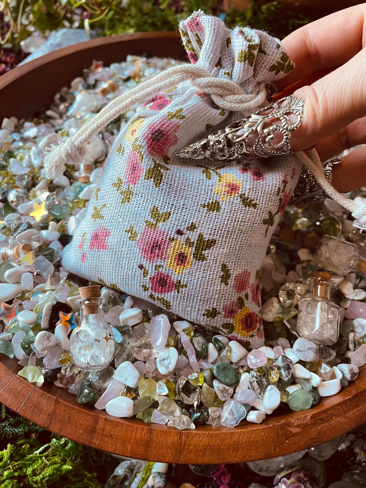 A small fabric drawstring pouch with a pink and yellow floral print, held by a hand over a wooden bowl filled with polished stones and small glass vials.