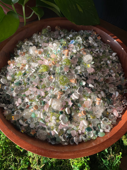 A wooden bowl filled with small decorative crystals photographed from above on a mossy surface with green leaves at the top.