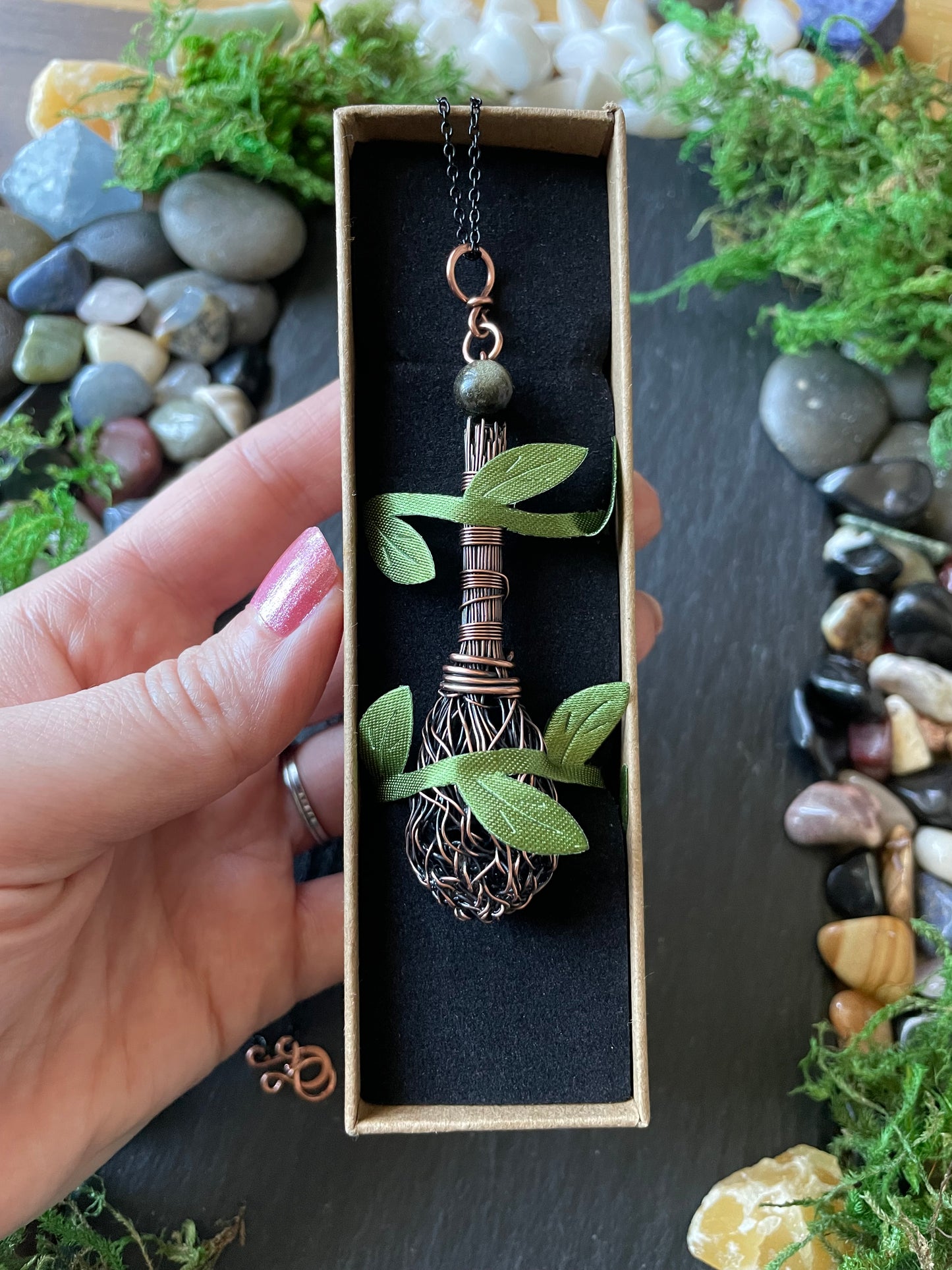 Hand holding a box with a decorative item and plants, surrounded by stones and moss.