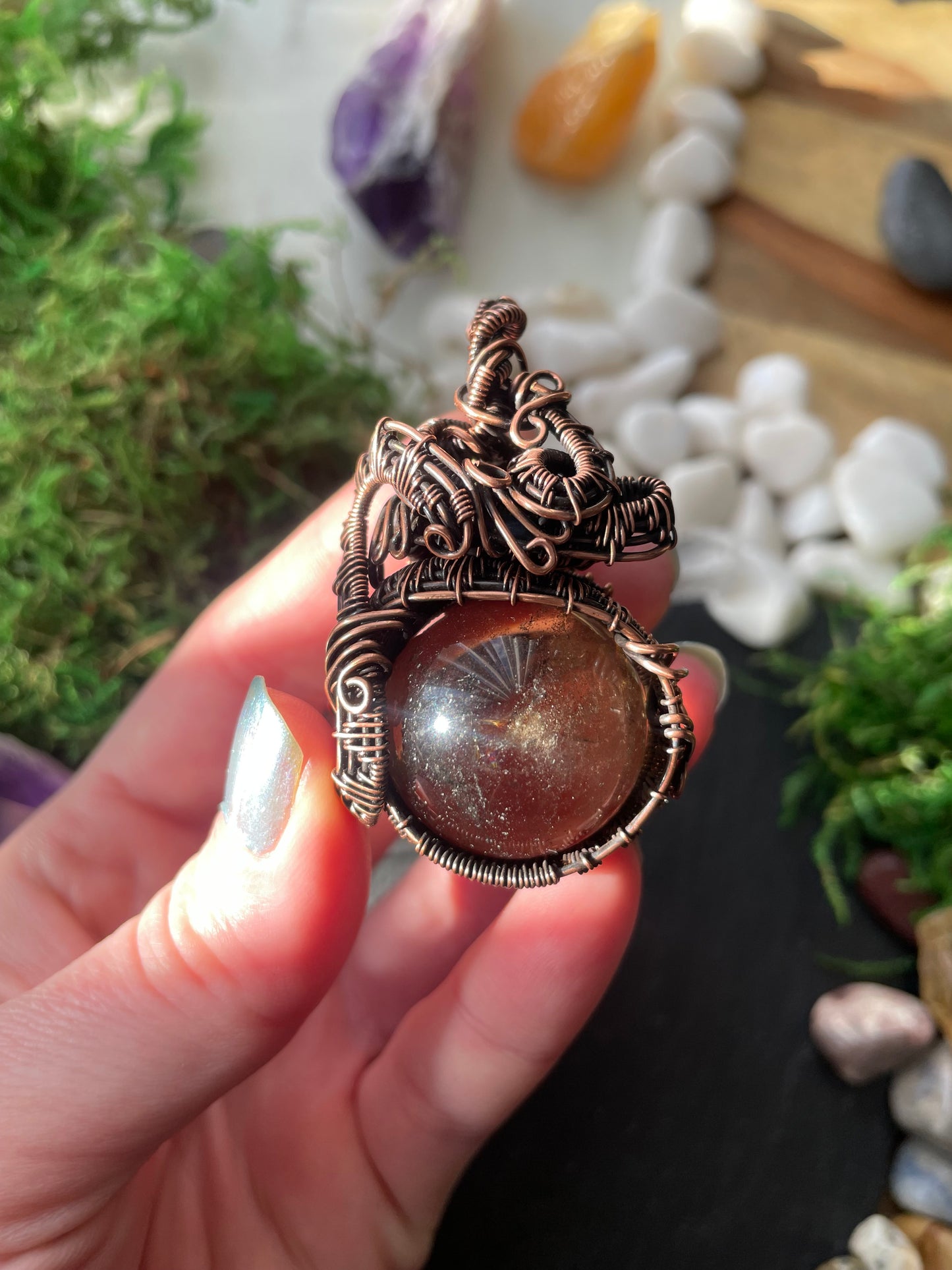 Hand holding a decorative amulet with smoky quartz crystal against a natural background with stones and plants.