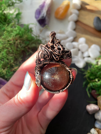 Hand holding a decorative amulet with smoky quartz crystal against a natural background with stones and plants.
