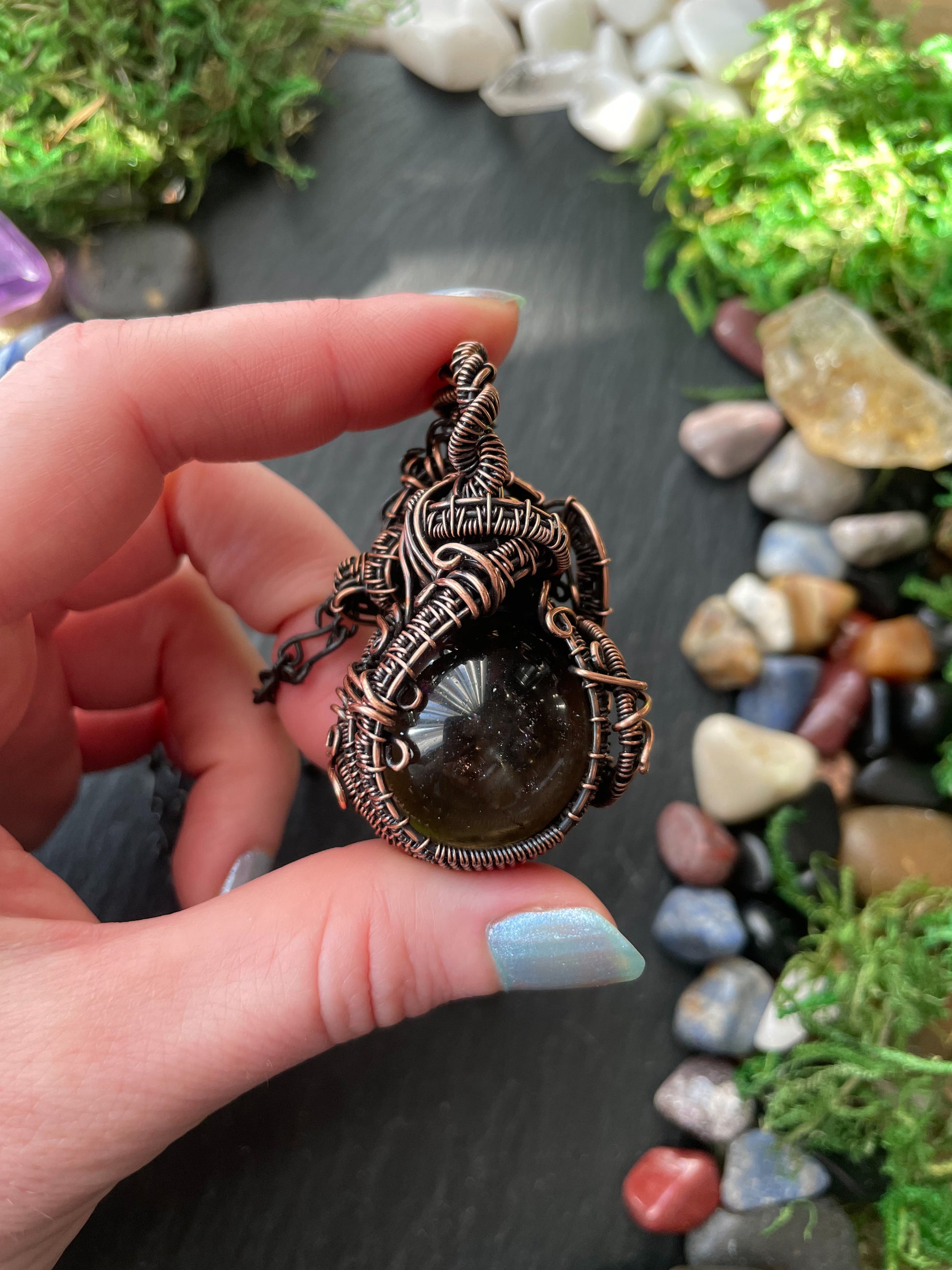 Hand holding a intricately designed copper amulet with a natural stone against a backdrop of stones and greenery.