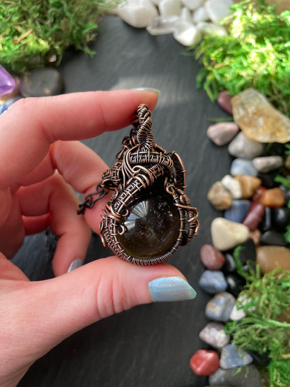 Hand holding a intricately designed copper amulet with a natural stone against a backdrop of stones and greenery.