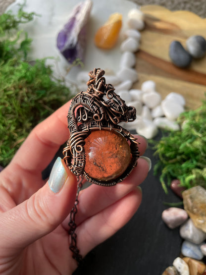 Hand holding a copper wire wrapped amulet with a large stone sphere against a natural background of stones and moss.
