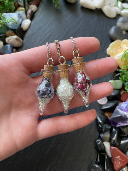 Hand holding mini vial charms with flowers and sparkles with a background of stones and crystals.