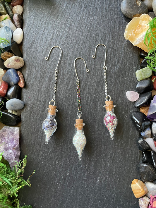Three glass vial charms on a dark stone surface with stones and plants around