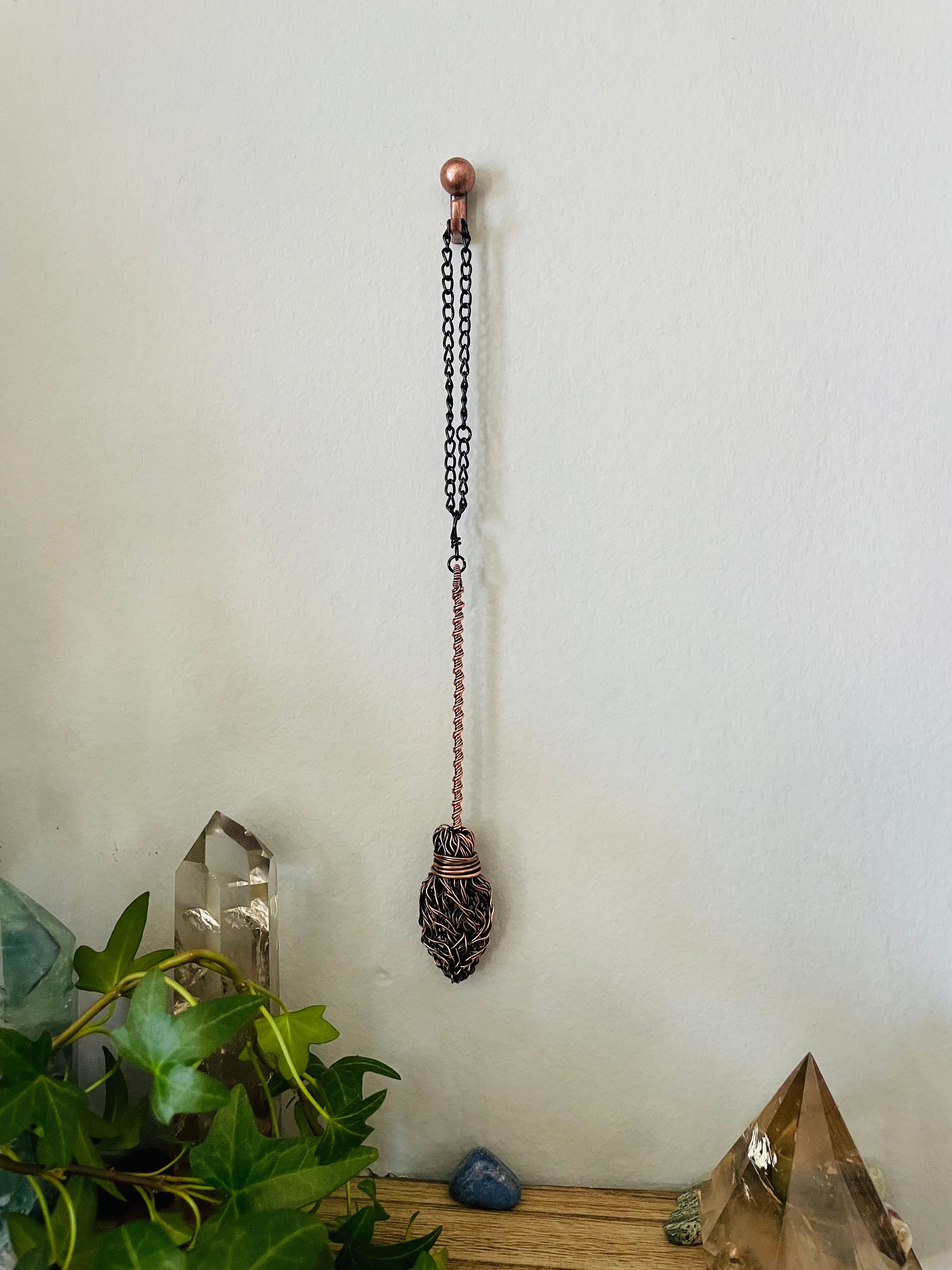 Decorative hanging broom pendant with chain on a white wall, next to crystals and plants.