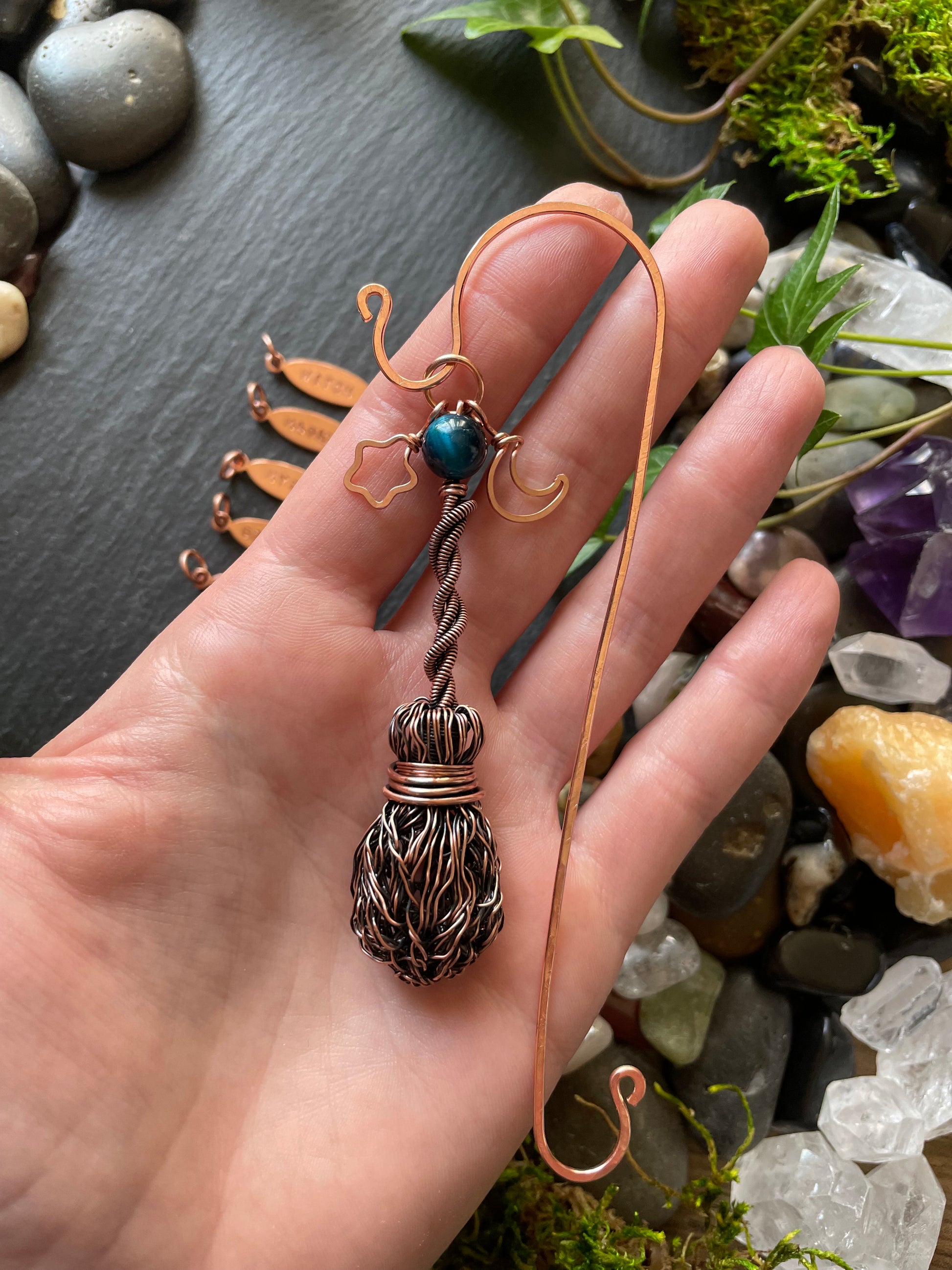 Hand holding a copper necklace with a decorative pendant against a natural stone and plant background