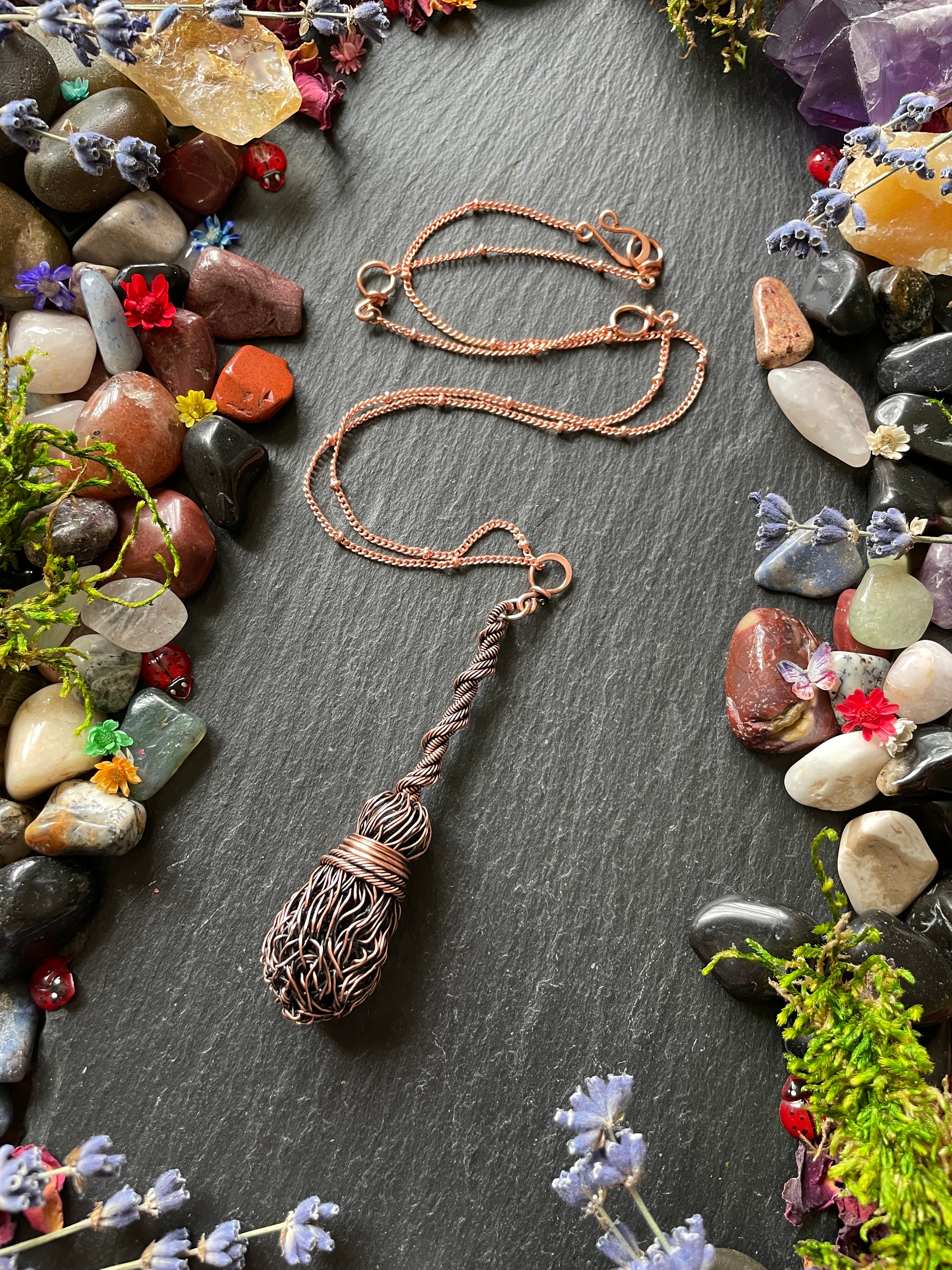 Antiqued copper miniature broom pendant with a twisted handle, surrounded by ladybugs and mini flowers, displayed on a stone slate
