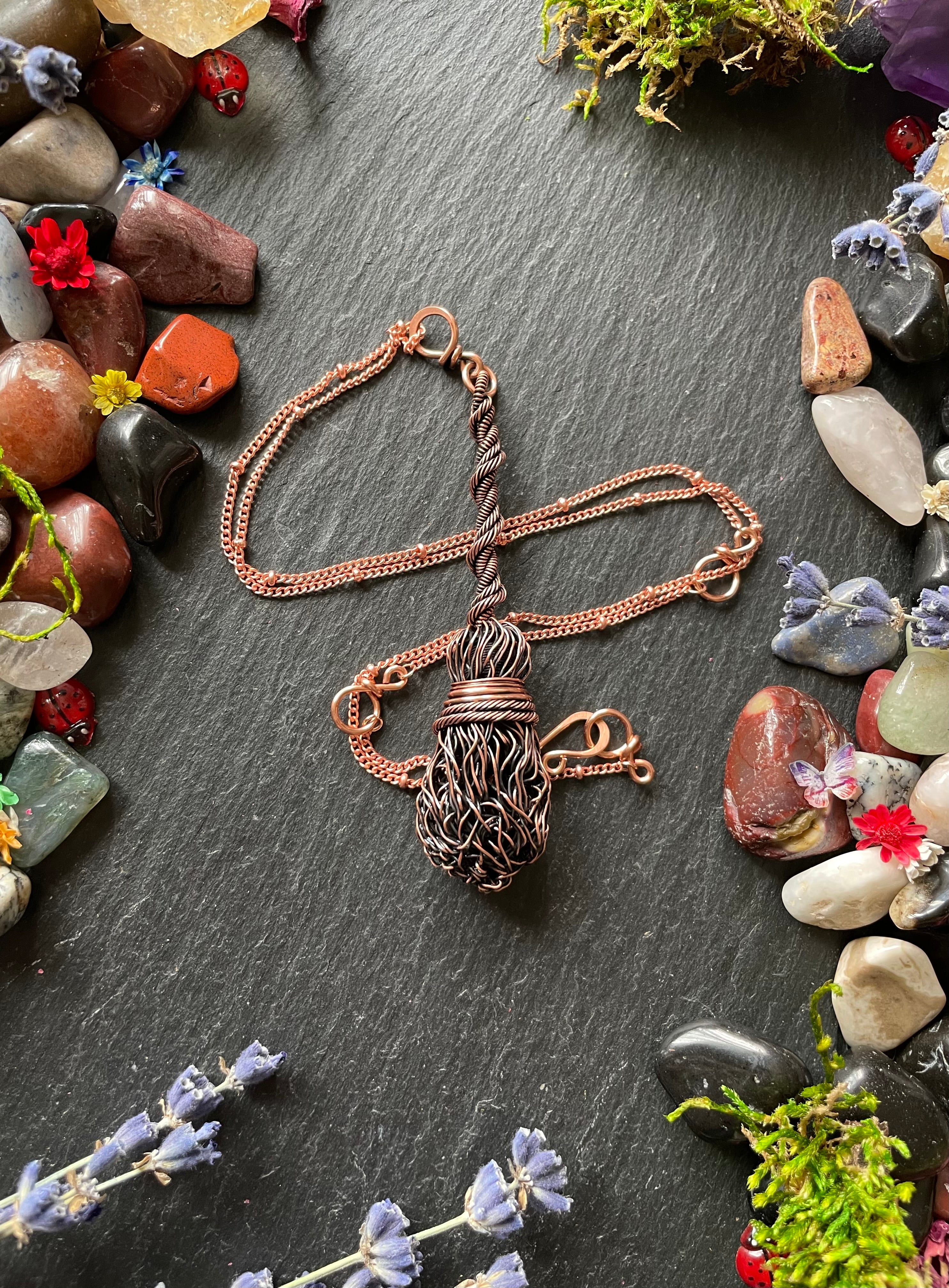 Antiqued copper miniature witch's broom pendant with a twisted rope handle, attached to a copper chain and surrounded by ladybugs and mini flowers and gemstones