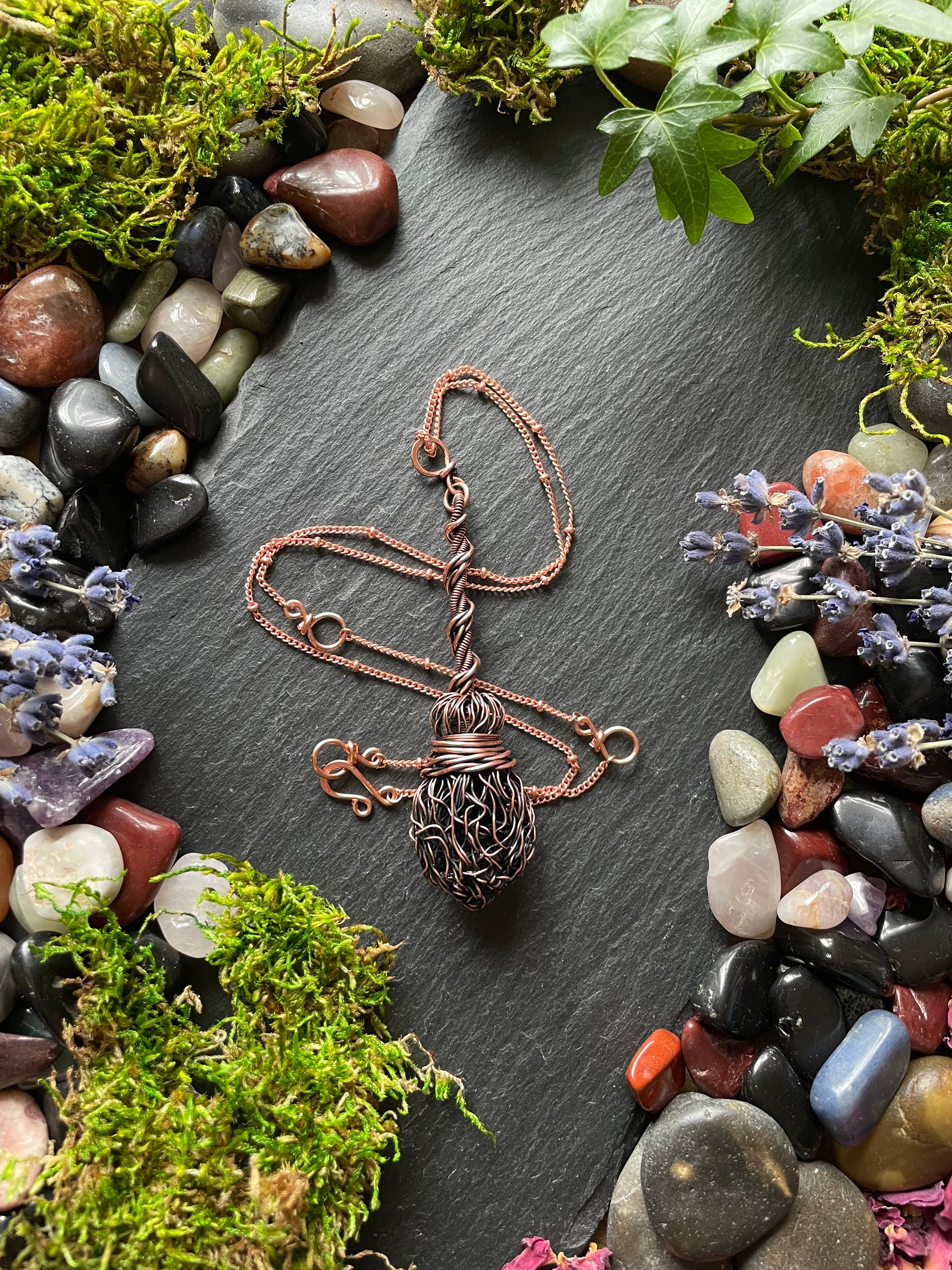 Miniature wire broom pendant attached to a copper chain, displayed on a stone slate surrounded by gems and foliage