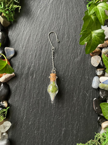 Glass vial charm with cork topper on a dark stone surface with greenery.