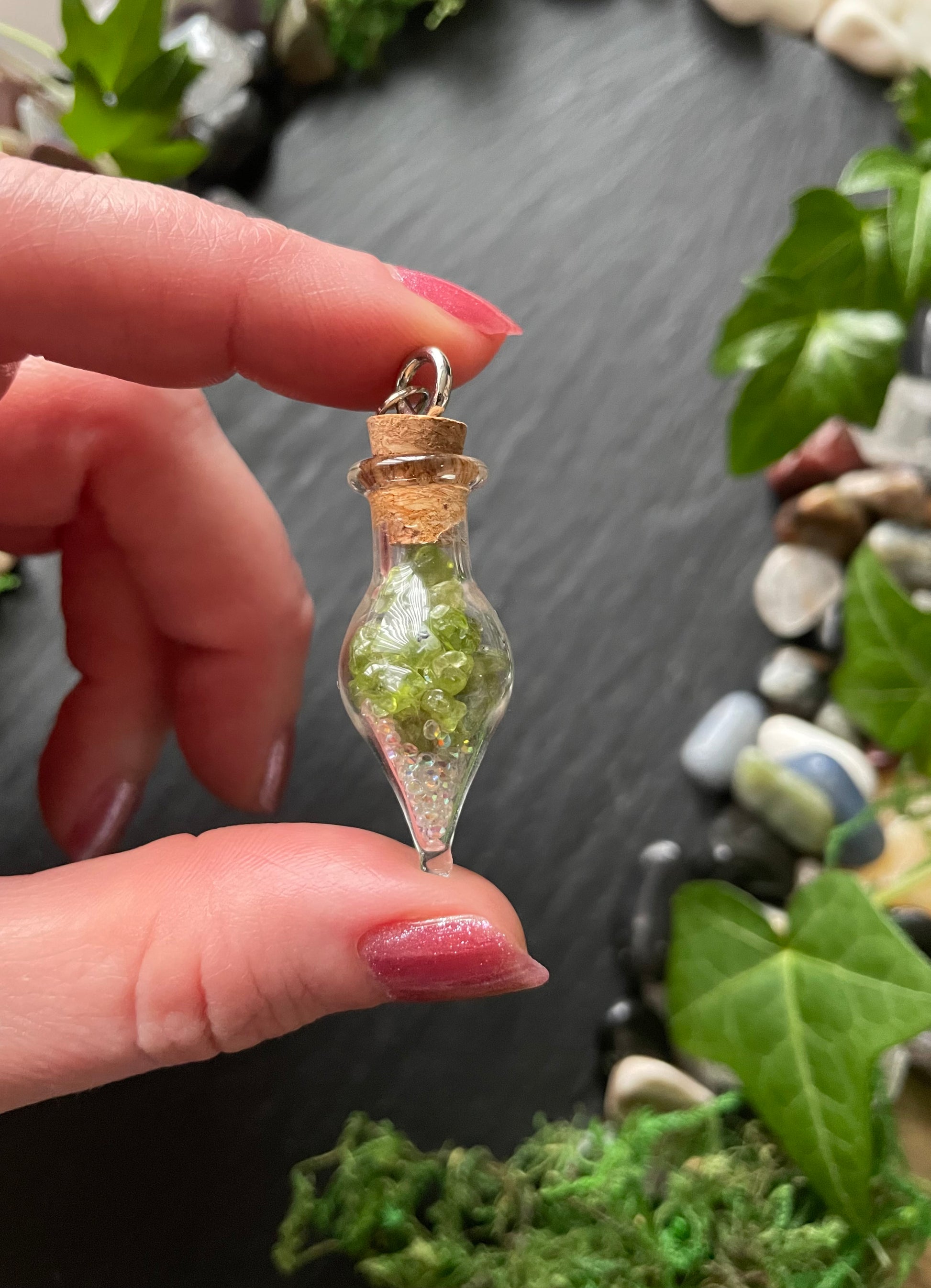 Hand holding a small glass bottle vial filled with green crystals, surrounded by stones and leaves.