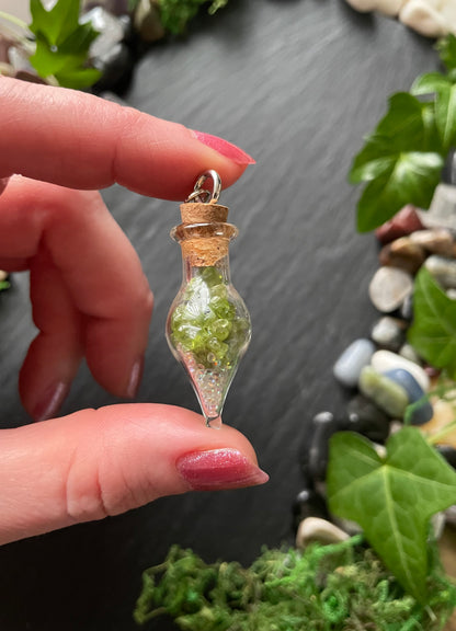 Hand holding a small glass bottle vial filled with green crystals, surrounded by stones and leaves.