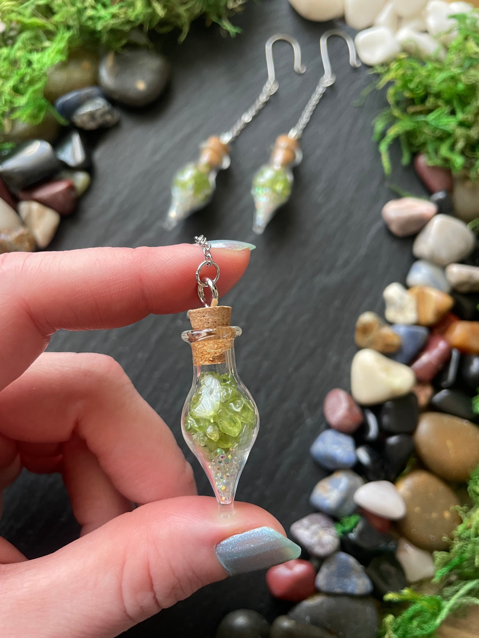 Hand holding a small glass bottle pendant with green crystals against a natural stone background