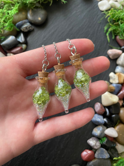Three small glass vial necklaces with green crystals held in a hand against a stone background.