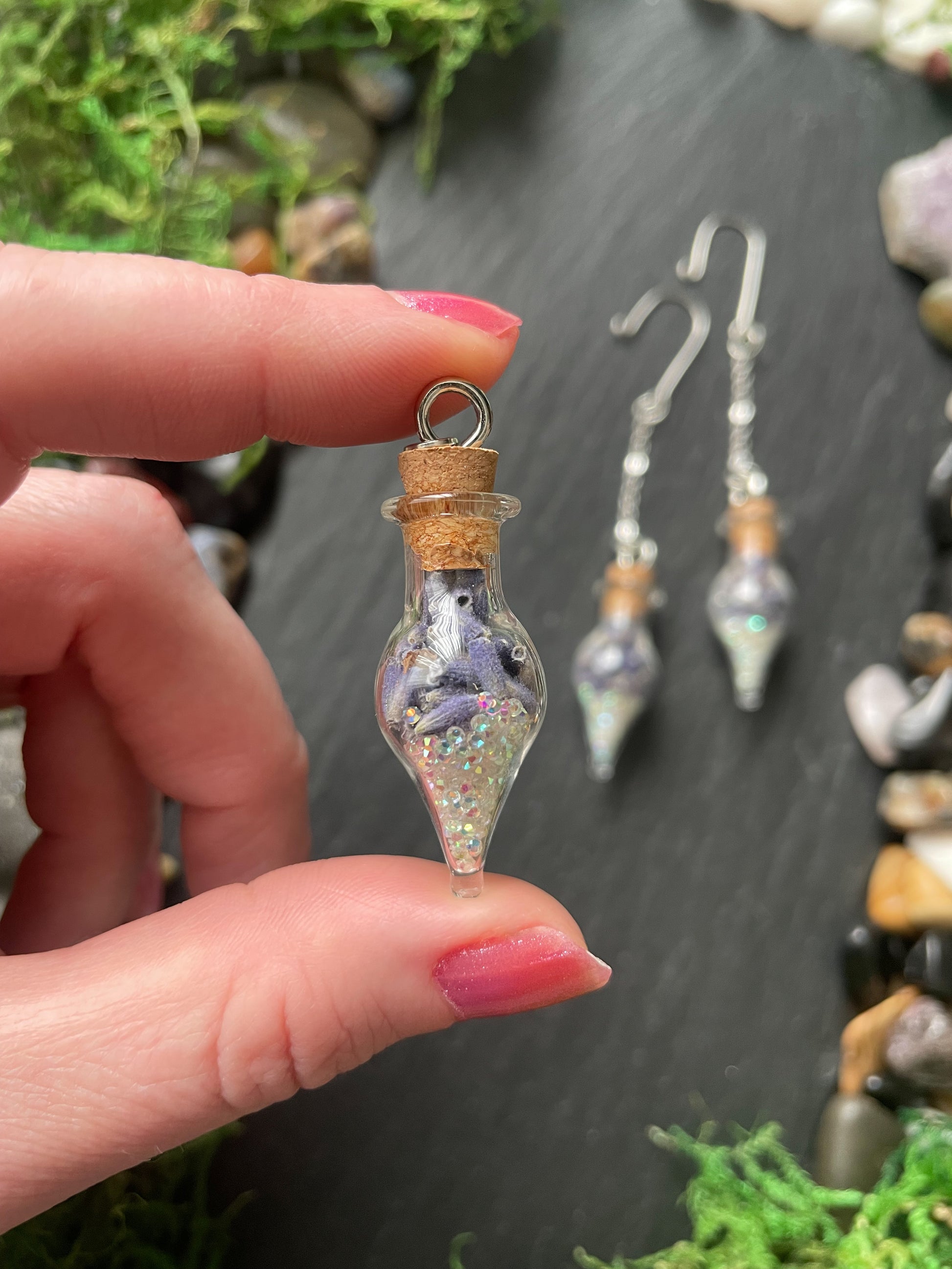 Hand holding a small glass vial with lavender flowers and glittery interior, against a natural background