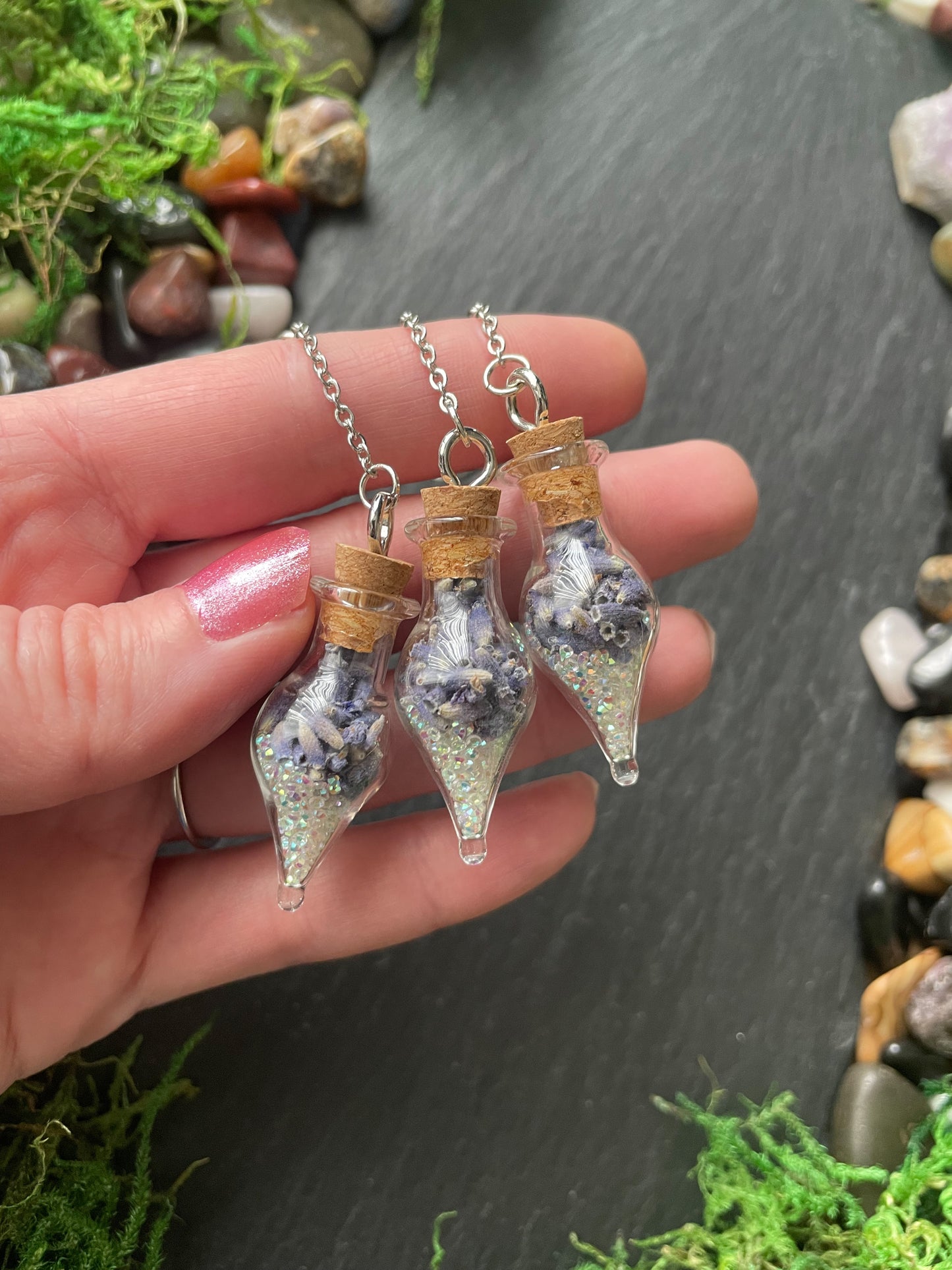 Three small glass vials with cork stoppers containing decorative elements, held by a hand against a natural stone background.