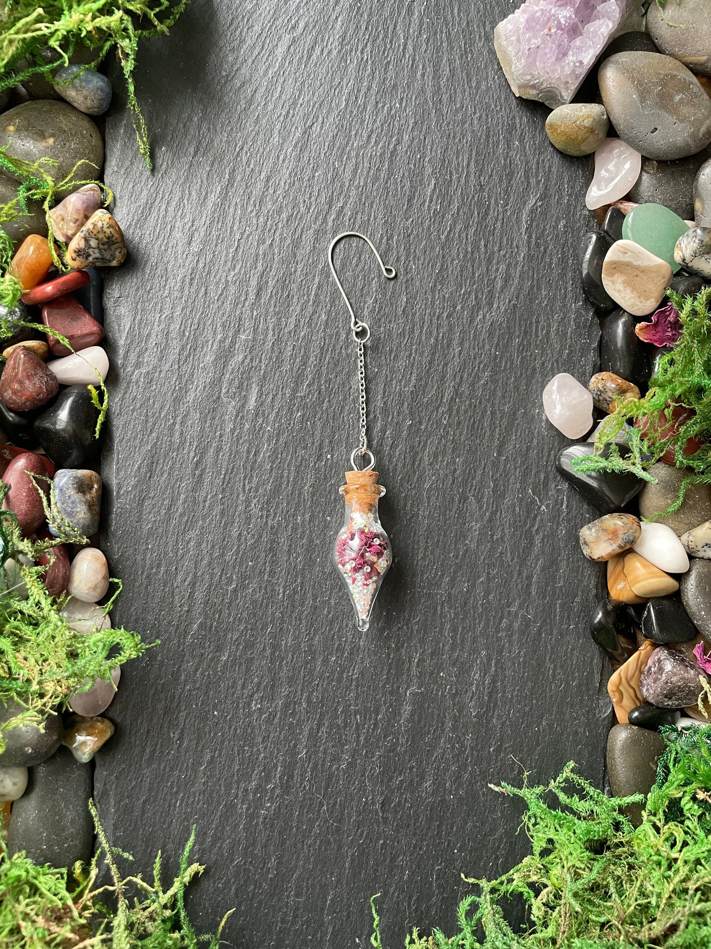 Small glass vials with rose petals and sparkles on a dark surface with stones and moss