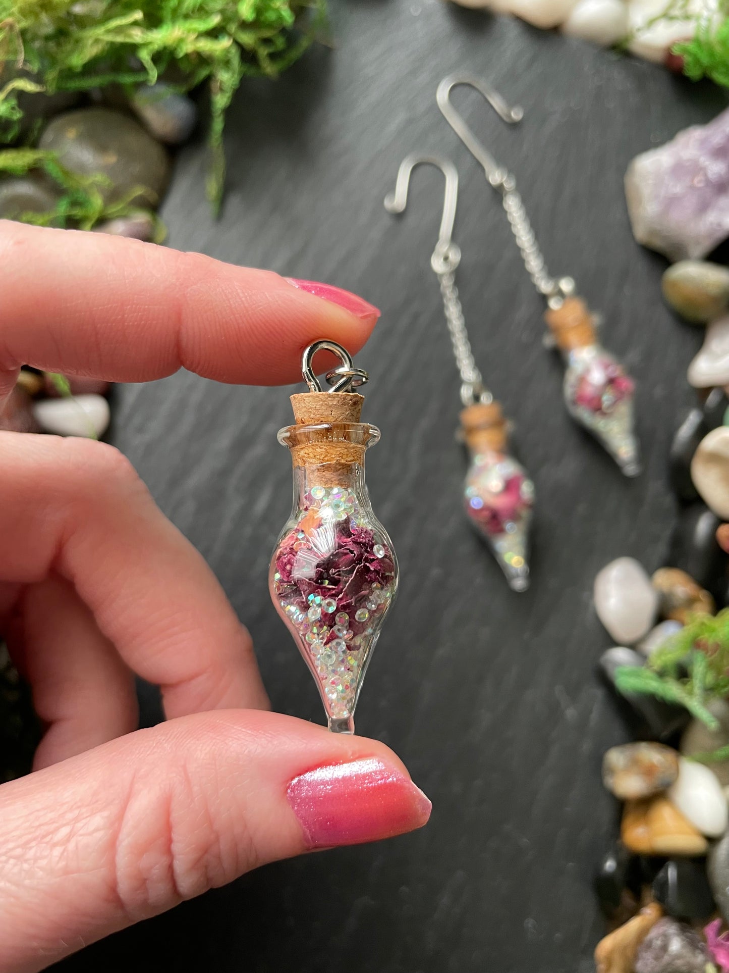 Hand holding a small glass bottle plant charms with decorative contents against a natural stone background.