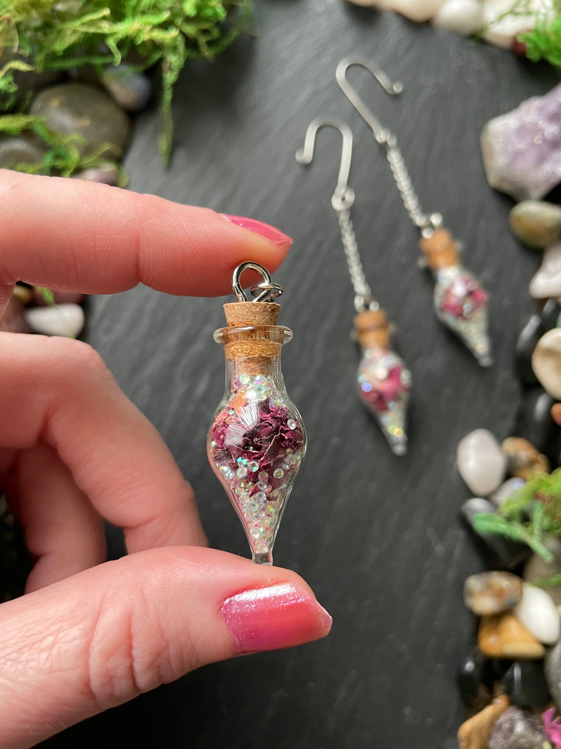 Hand holding a small glass bottle plant charms with decorative contents against a natural stone background.