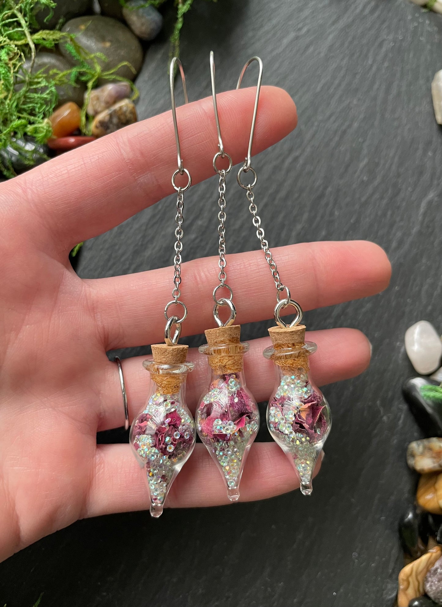 Hand holding two decorative vials filled with flowers on a dark surface.