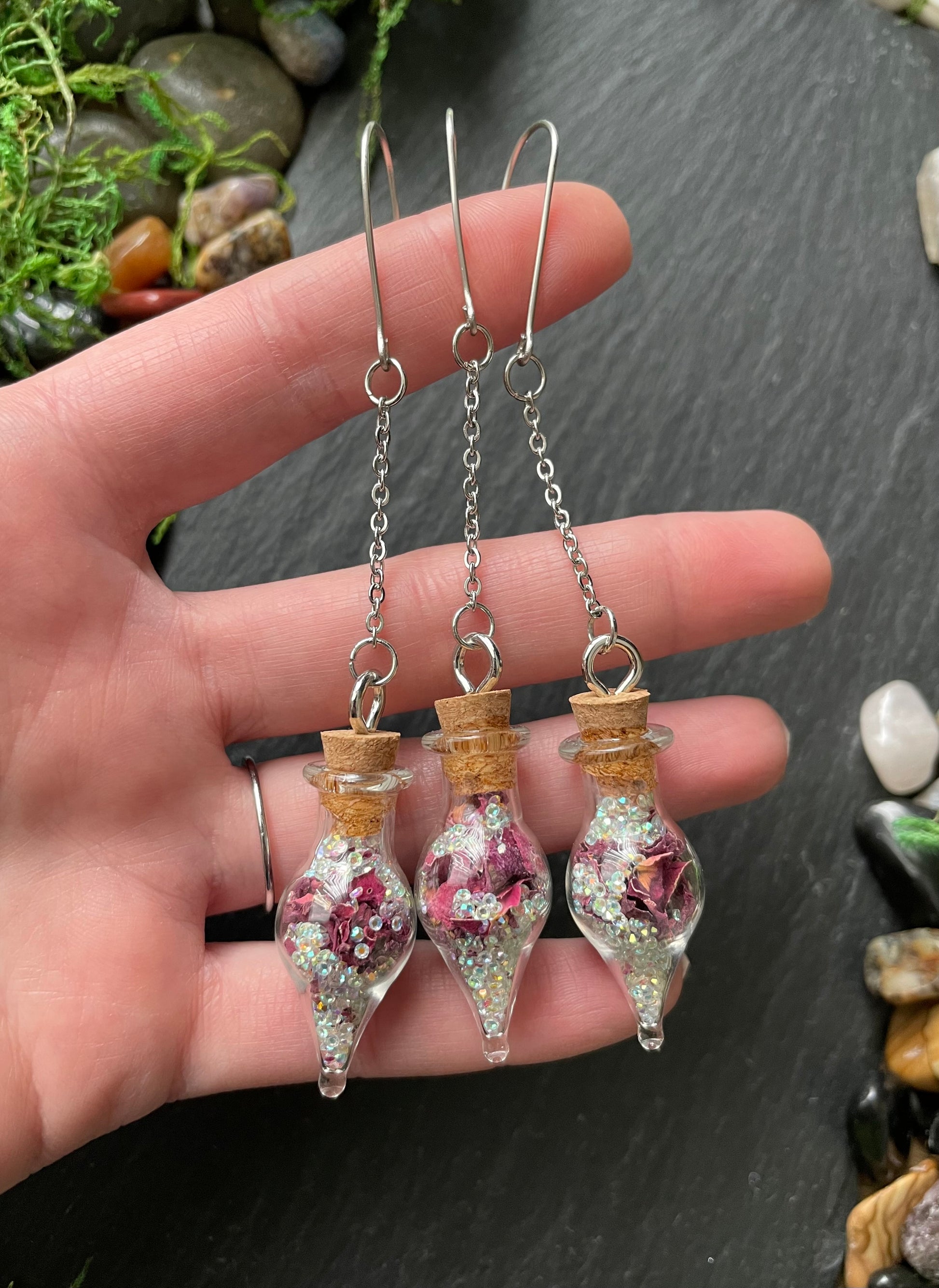 Hand holding two decorative vials filled with flowers on a dark surface.