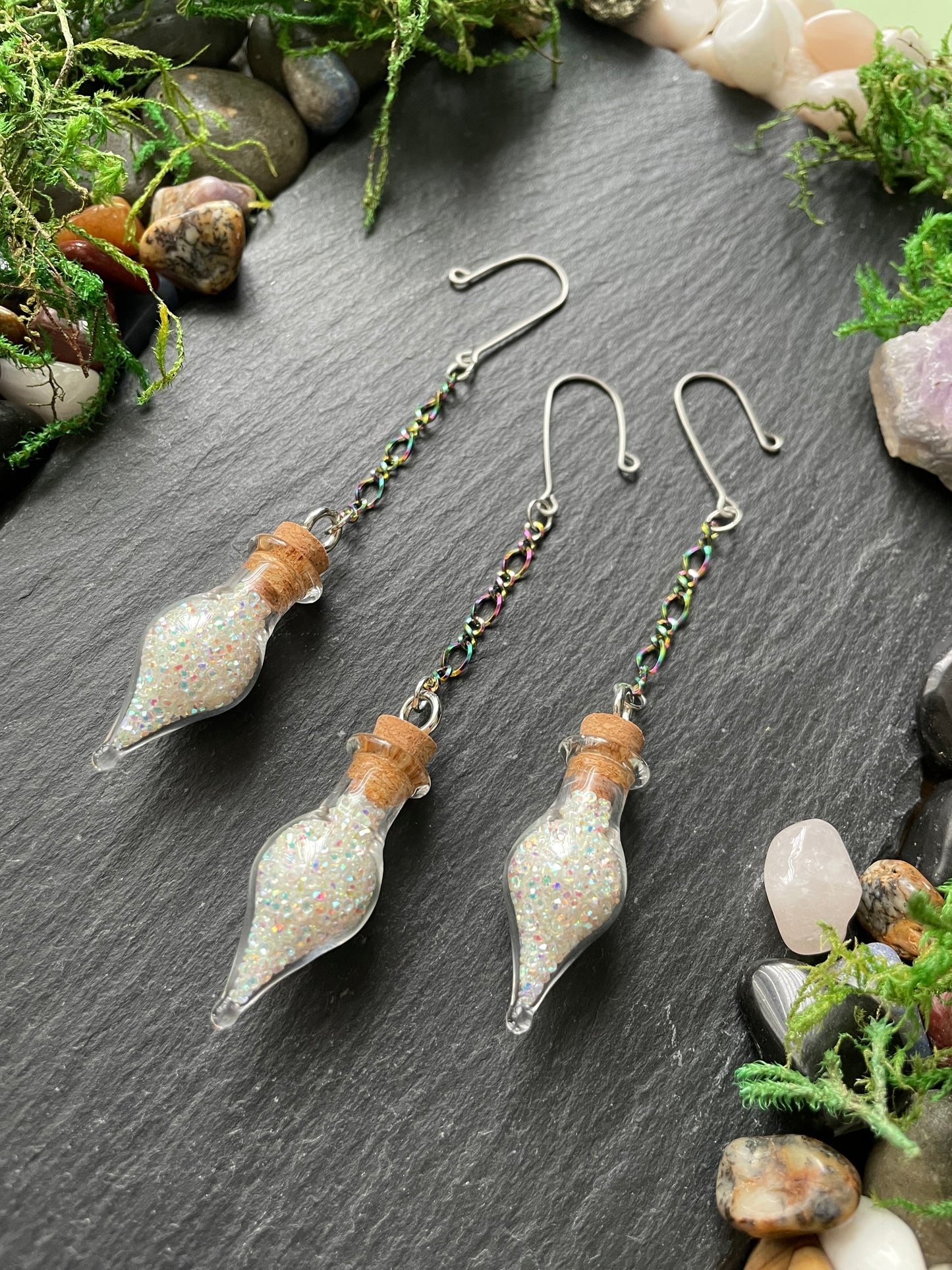 Mini glass vials with cork stoppers on a dark surface surrounded by greenery and stones.