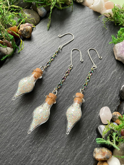 Mini glass vials with cork stoppers on a dark surface surrounded by greenery and stones.